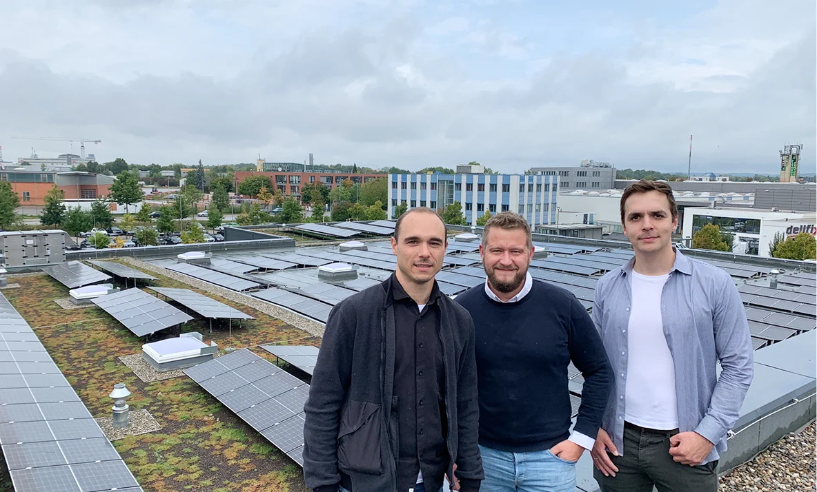Drei Personen stehen auf einem Gebäudedach vor einer Photovoltaikanlage mit Solarmodulen in einer städtischen Umgebung