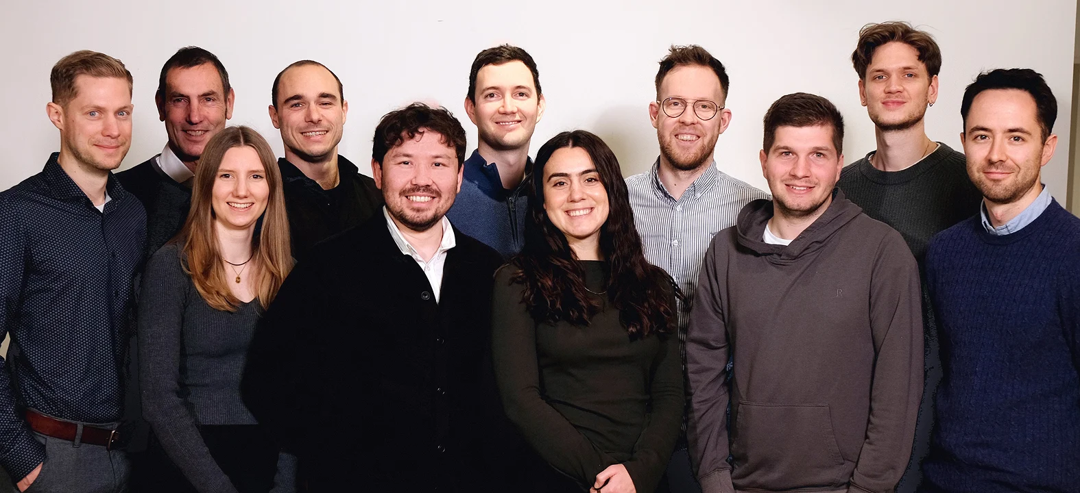 Mehrere Teammitglieder stehen dicht nebeneinander vor einer hellen Wand und blicken in die Kamera