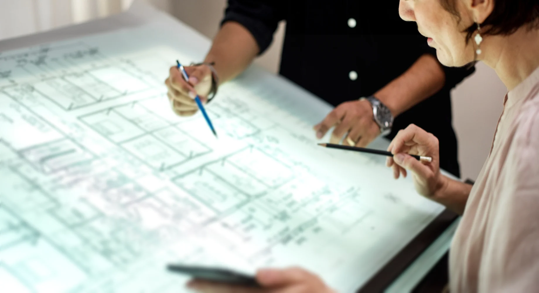 Zwei Personen prüfen einen beleuchteten Bauplan auf einem Tisch und zeigen mit Stiften auf technische Zeichnungen eines Grundrisses